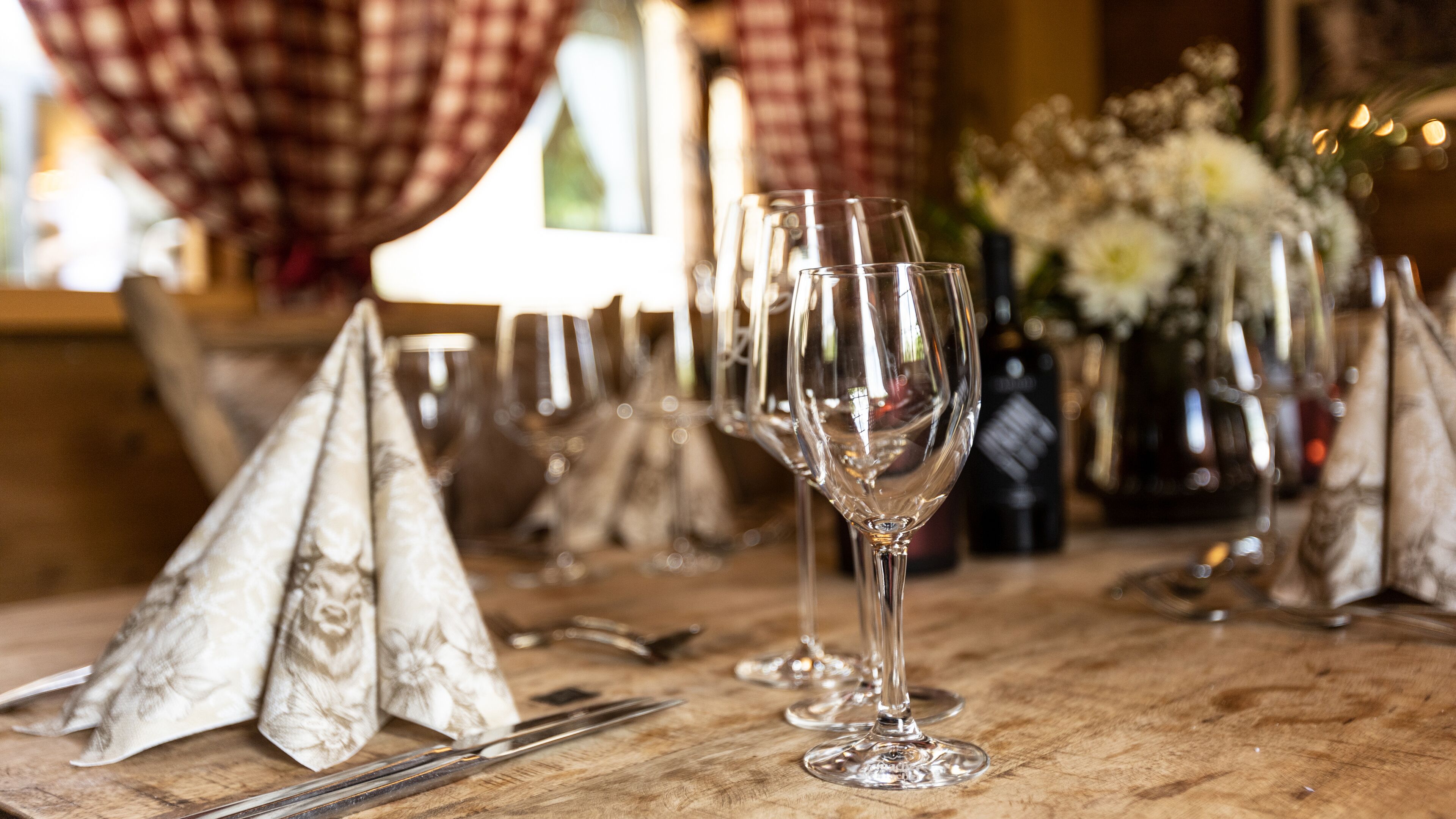 Una mesa de comedor con copas de vino y cubiertos, un jarrón con flores, y cortinas a cuadros al fondo.