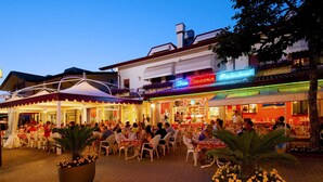 An outdoor dining area with tables and chairs, a neon sign for a restaurant, and a building with balconies.