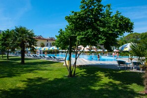 A swimming pool area with lounge chairs and umbrellas, surrounded by trees and buildings in the background. - Camping Parco Capraro (Jesolo)