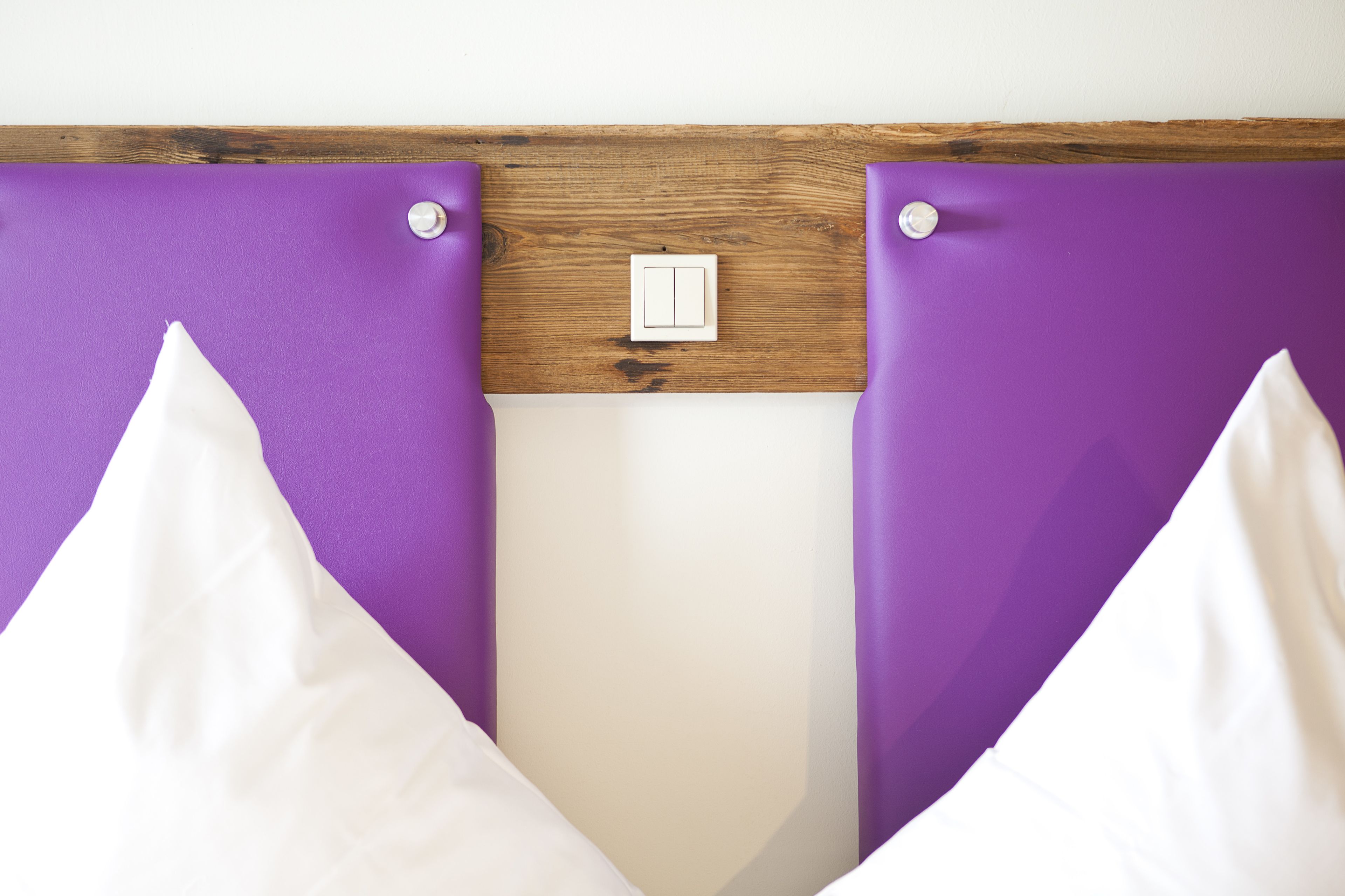 A bed with purple headboard and white pillows, a wooden headboard, and a light switch on the wall.