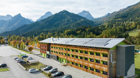 A modern hotel with a green facade and solar panels, surrounded by a parking lot and autumn trees, with mountains in the background.