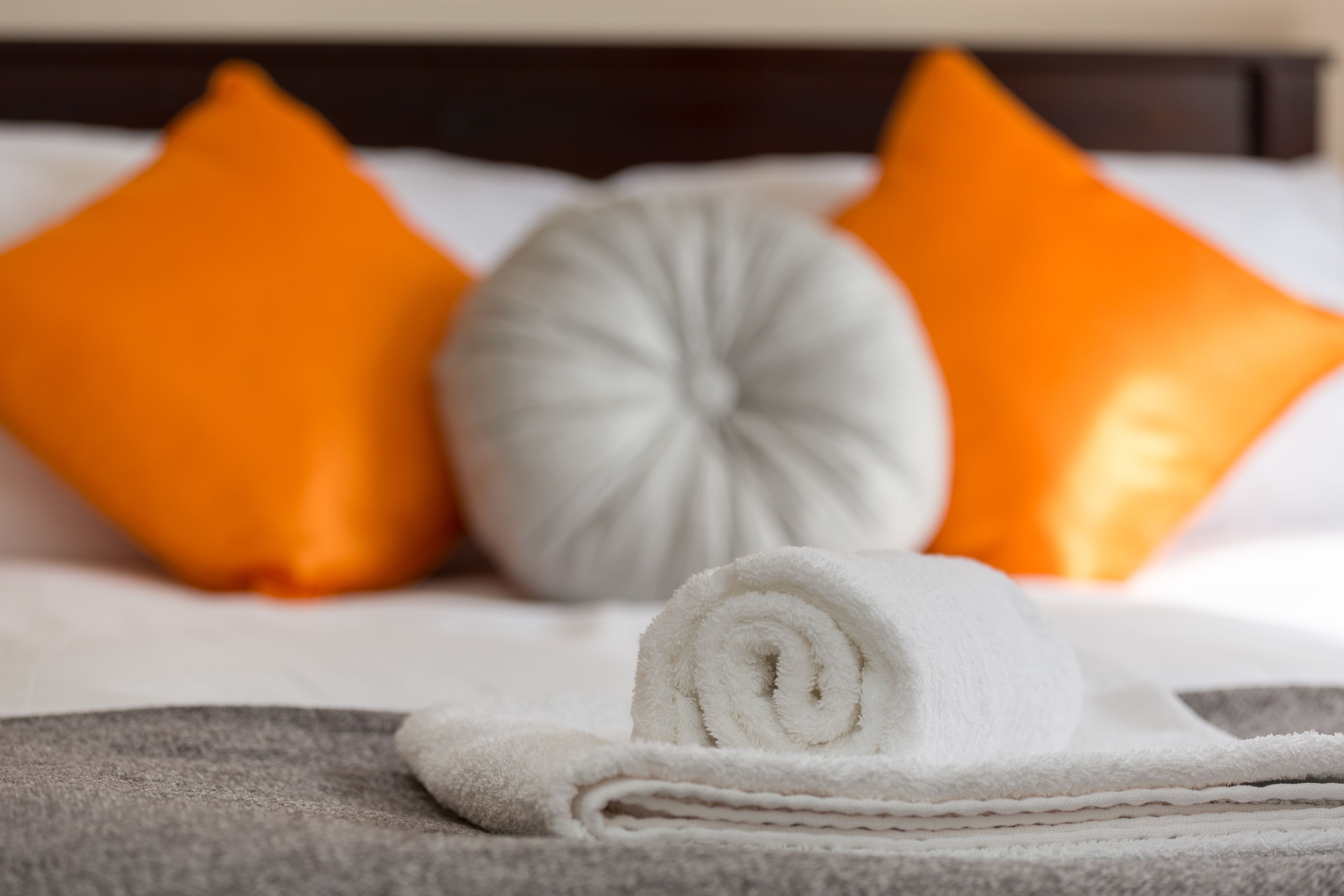 A neatly made bed with a rolled white towel, two orange pillows, and a gray pillow.