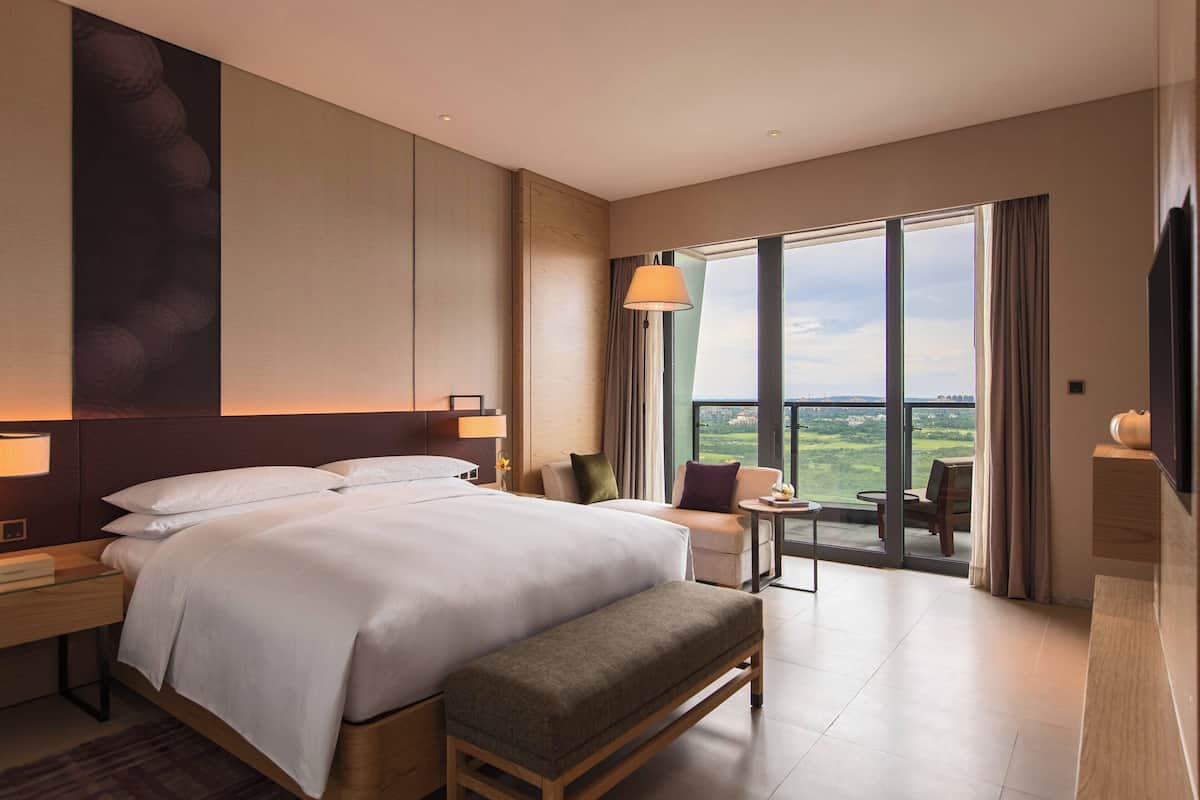 Habitación de hotel con una cama grande, un banco, una mesita, un sofá y vistas al exterior.