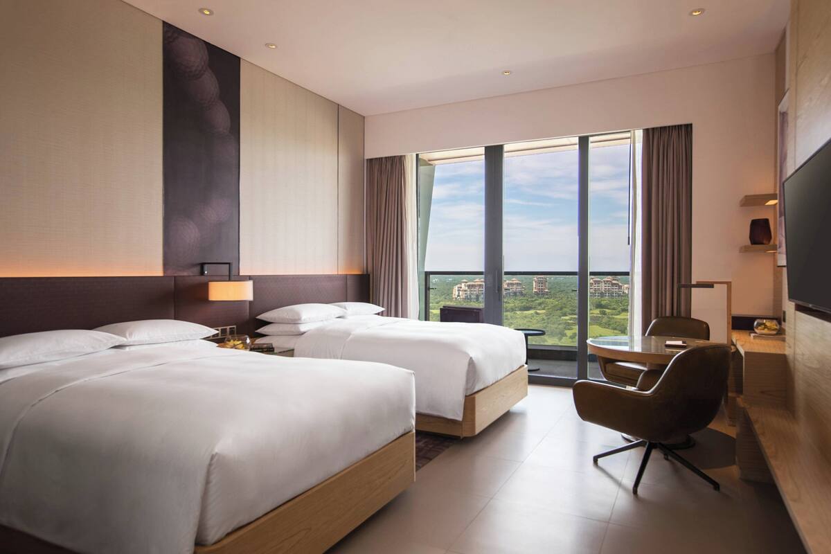 Habitación de hotel con dos camas, un escritorio, una silla y un ventanal con vistas a la ciudad.