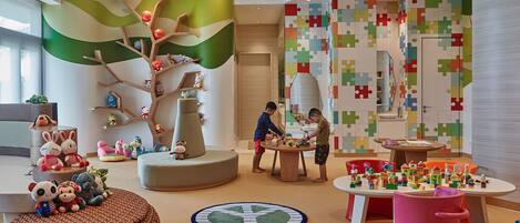 Un espace de jeux pour enfants avec une grande fresque représentant un arbre, un mur de jeux de puzzle et divers jouets et tables d’activités.