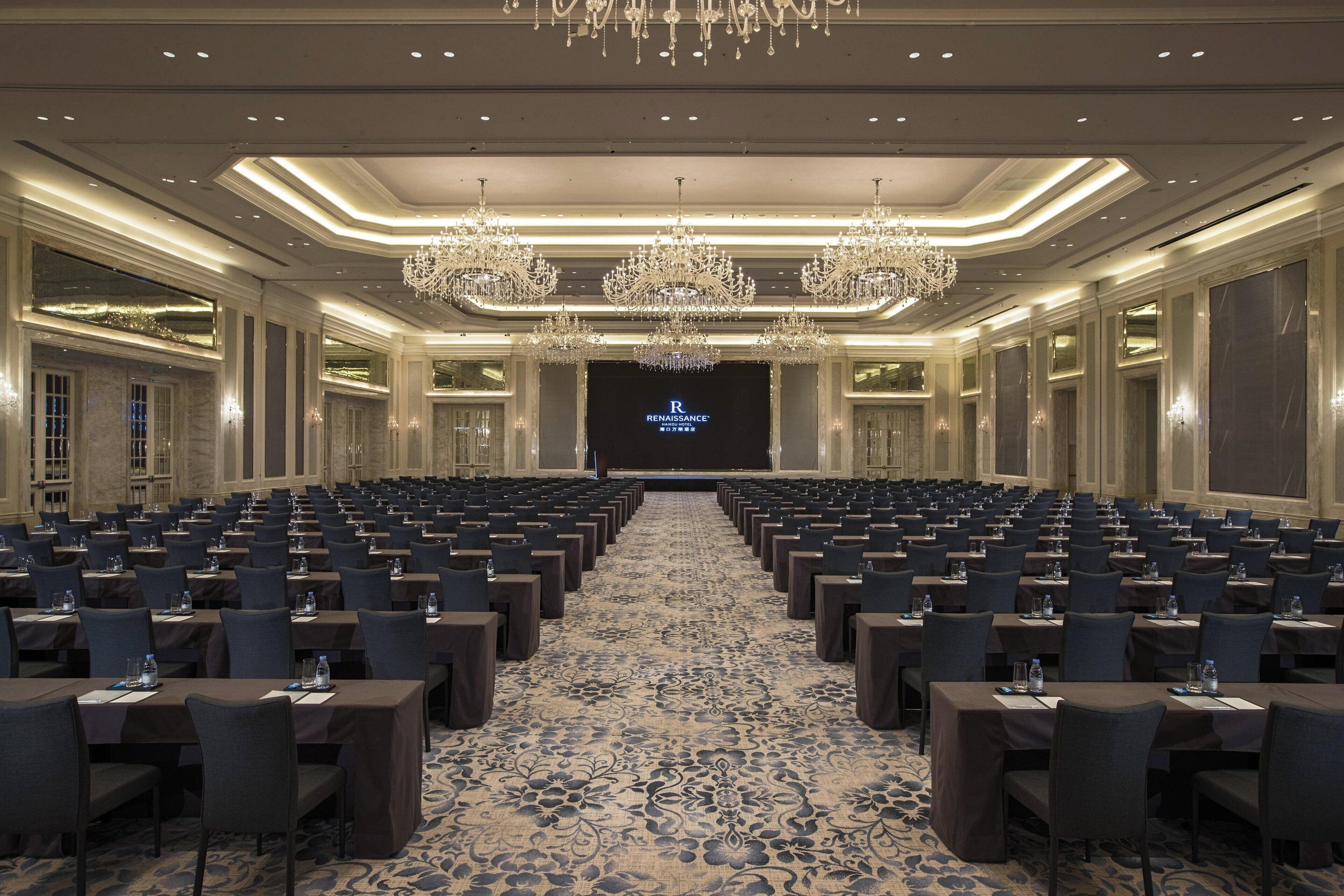 A conference room with rows of tables and chairs, a large screen at the front, and chandeliers on the ceiling.