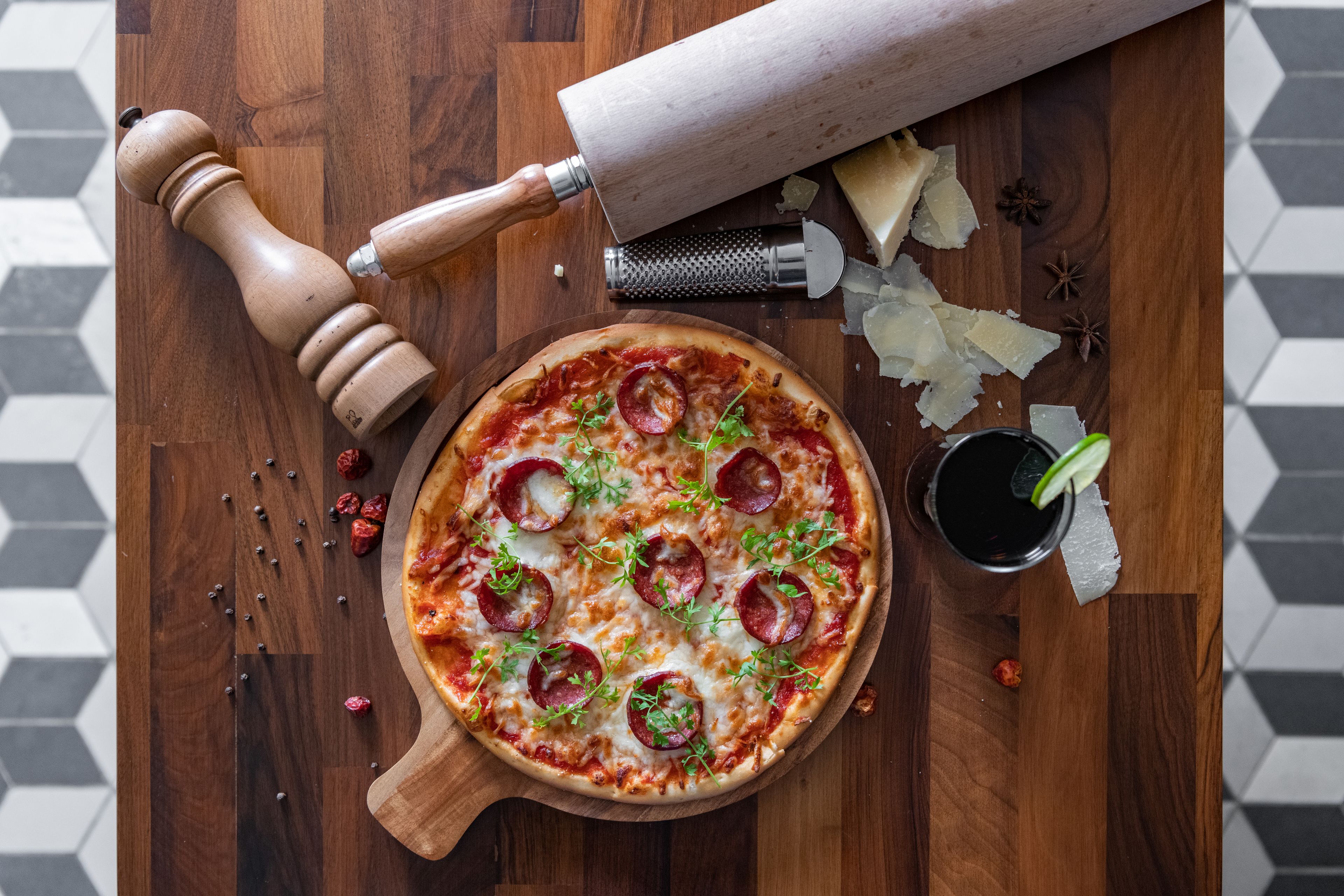 A pizza on a wooden board with pepper mill, grater, and star anise.