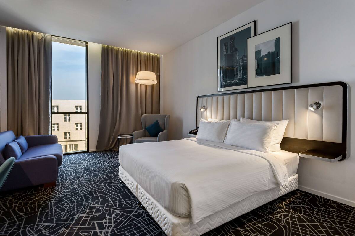 Une chambre d’hôtel avec un grand lit, un canapé, un fauteuil et une vue sur la ville par la fenêtre.