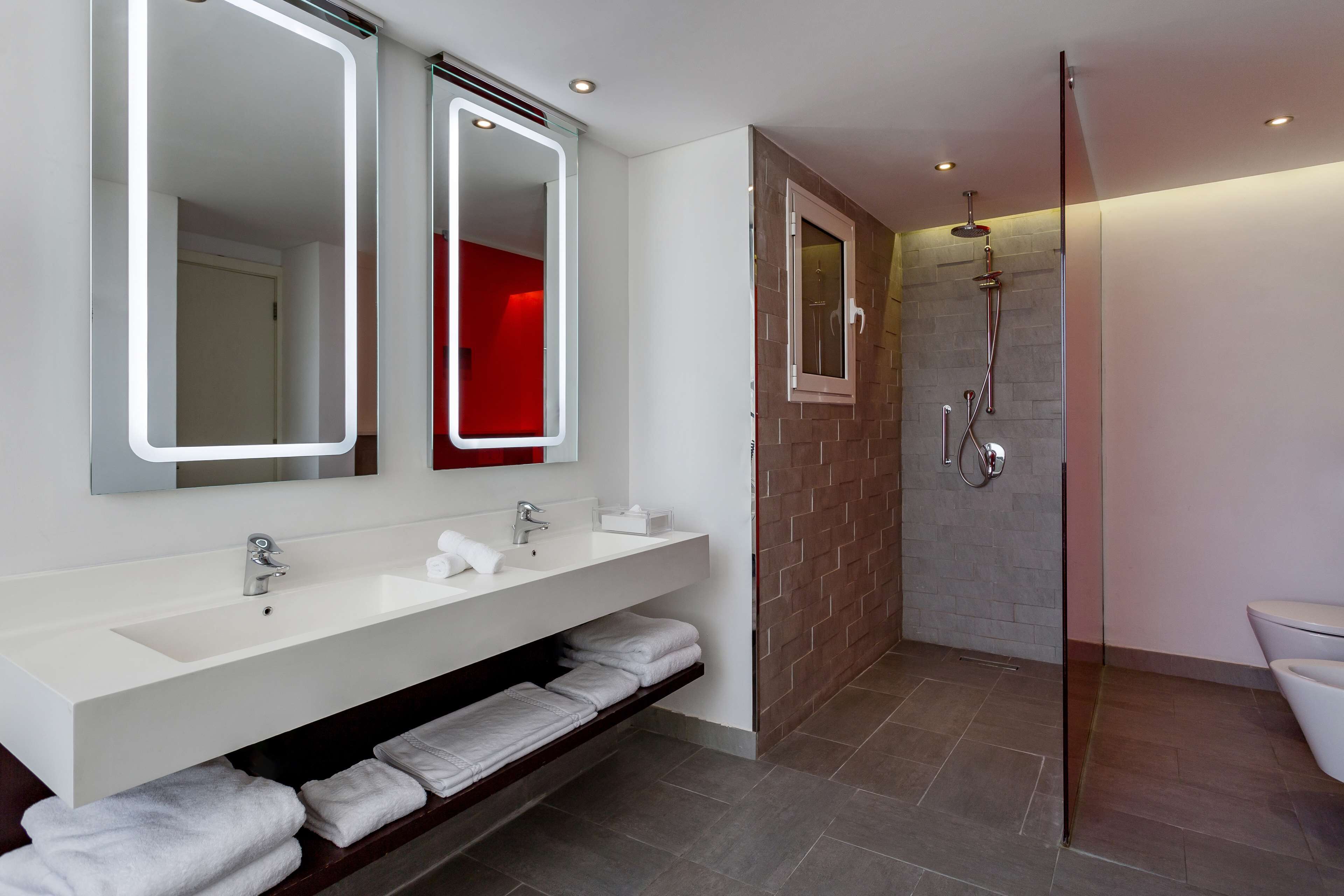 A modern bathroom with a white vanity, two mirrors, a glass-enclosed shower, and a red accent wall.