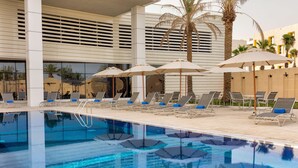 Piscina con sillas de descanso y sombrillas, rodeada de palmeras y arquitectura moderna.