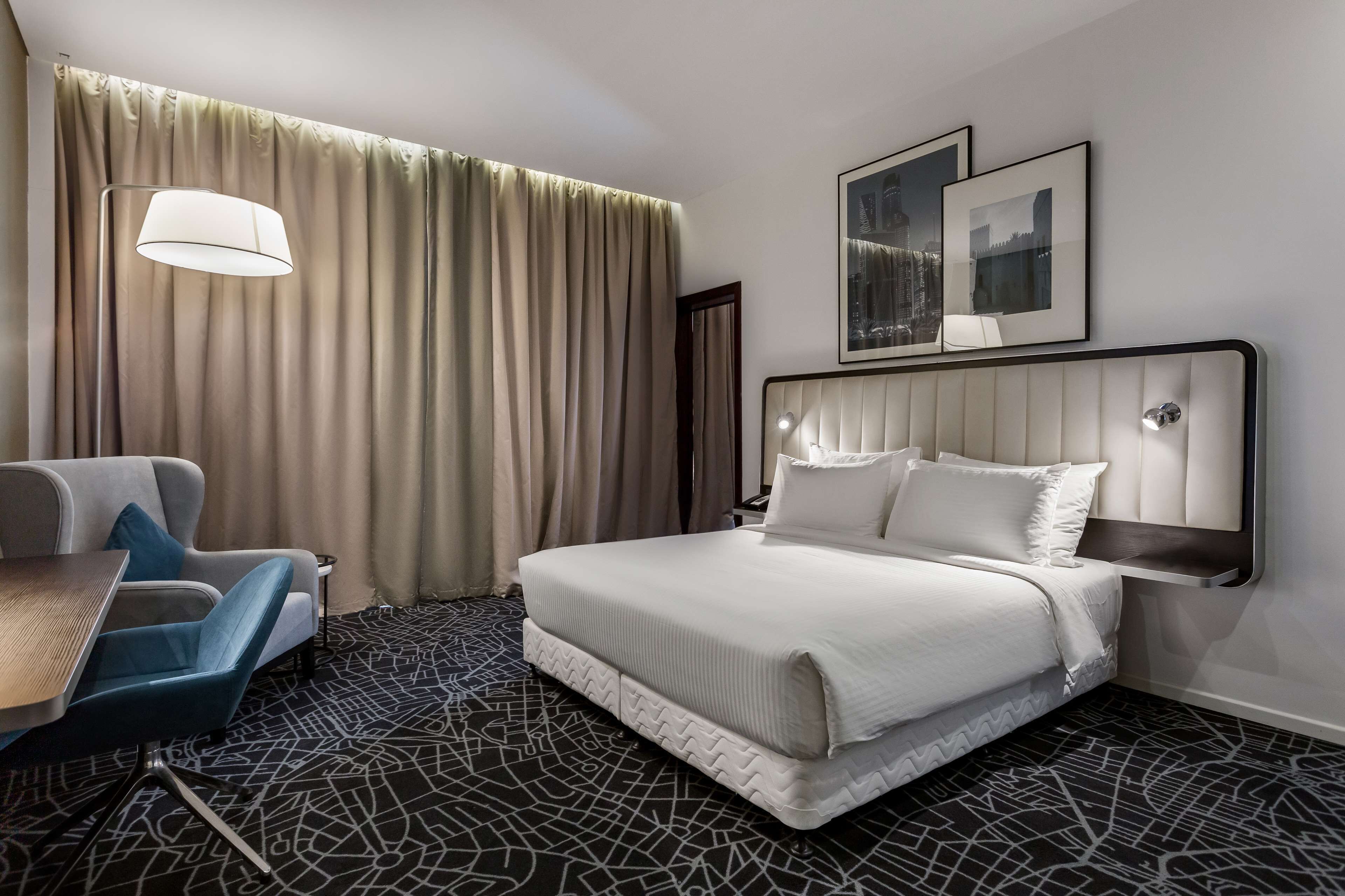 Ein Hotelzimmer mit Bett, Schreibtisch, Stuhl, Lampe und einem großen Fenster mit Vorhängen.
