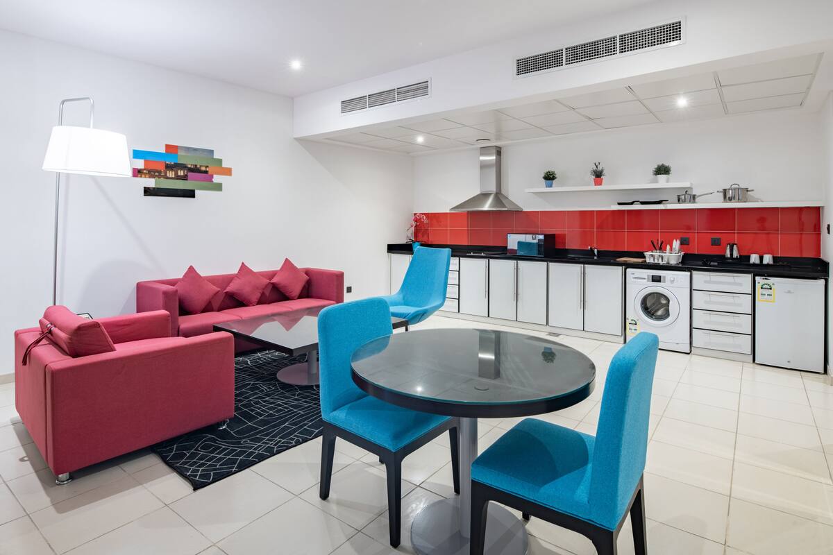 Un salon moderne avec un canapé rouge, des fauteuils bleus et une table en verre. On aperçoit, au fond, une cuisine avec des carreaux rouges et des meubles blancs.