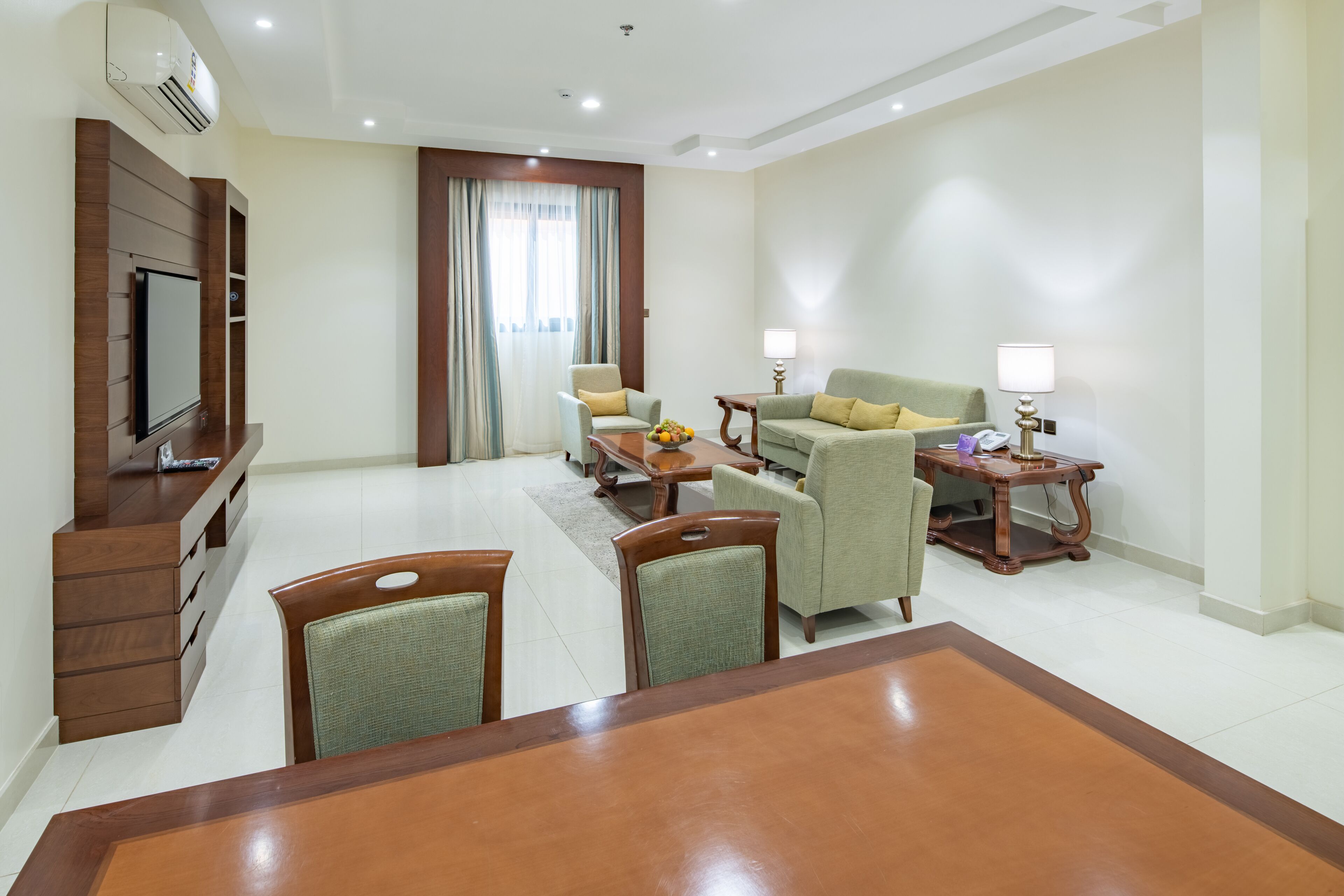 A hotel room with a wooden dining table, two armchairs, a sofa, a TV mounted on a wooden panel, and a small side table with a lamp.