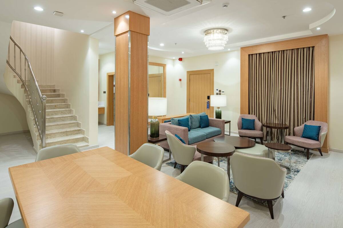 A hotel lobby with a wooden staircase, a seating area with sofas and armchairs, a dining table, and a chandelier.