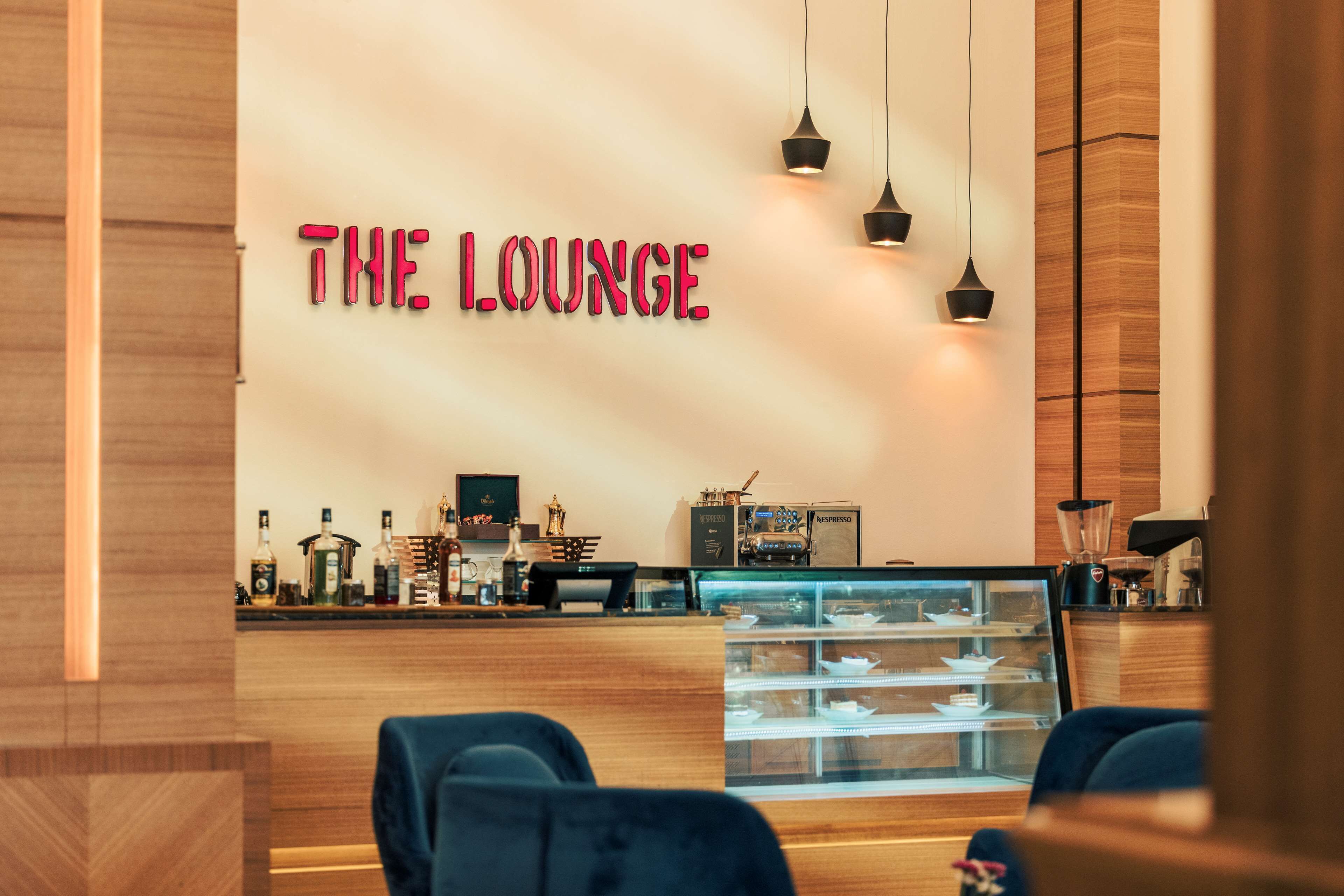 A modern lounge with a wooden reception desk, a glass display case, and a neon sign reading "THE LOUNGE".
