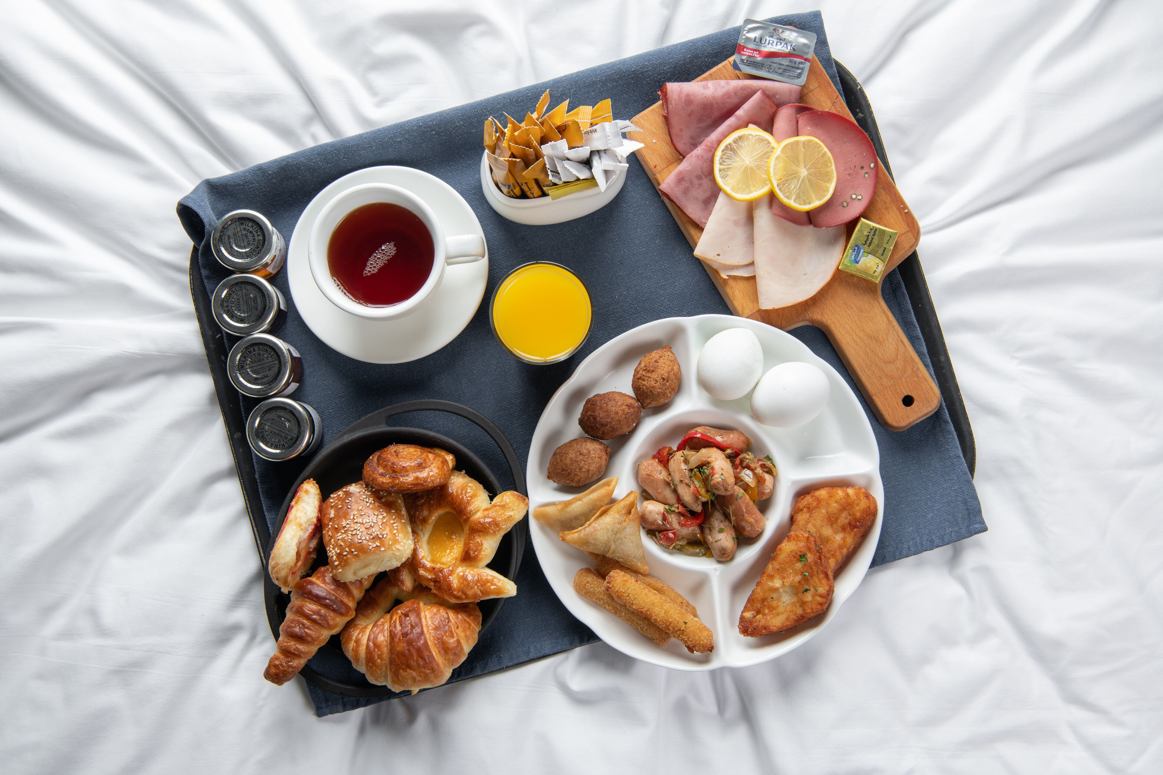 Un vassoio con prodotti per la colazione, tra cui croissant, uova, salsiccia e prosciutto, su un letto.