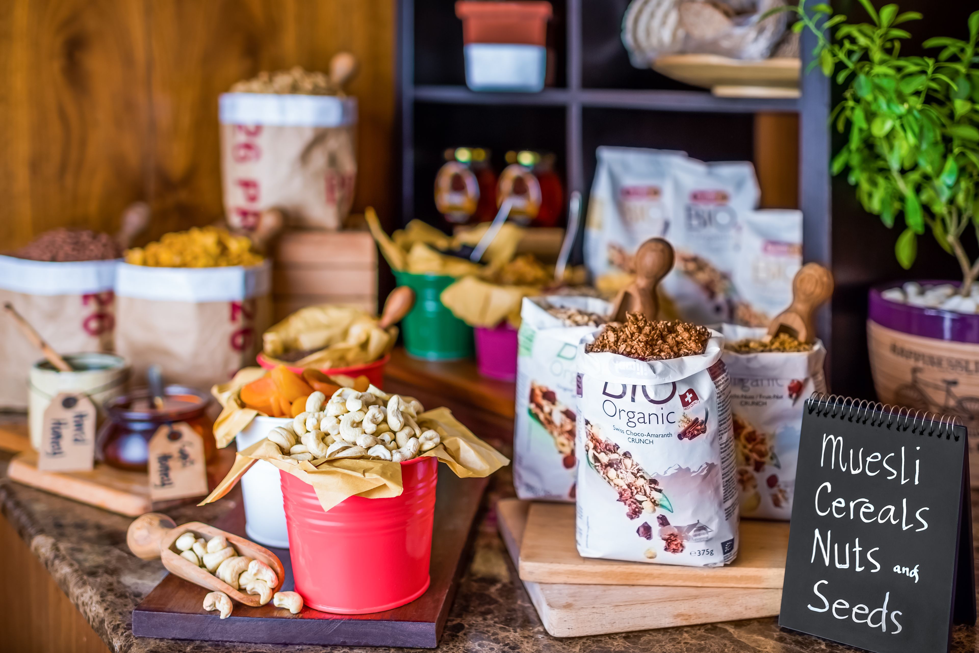 Keittiön työtasolla on erilaisia elintarvikkeita ja säilytysastioita, kuten punainen kuppi, joka on täynnä pähkinöitä, valkoinen pussi, jossa on merkintä ”DIY Organic Crunch”, ja pieni taulu, johon on kirjoitettu teksti ”Muesli Cereals Nuts and Seeds”.