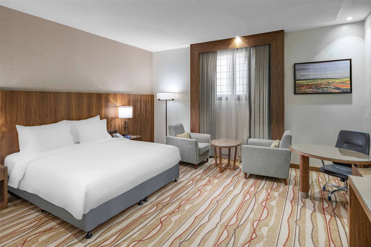 A hotel room with a large bed, a desk with a chair, two armchairs, a small table, a lamp, and a painting on the wall.