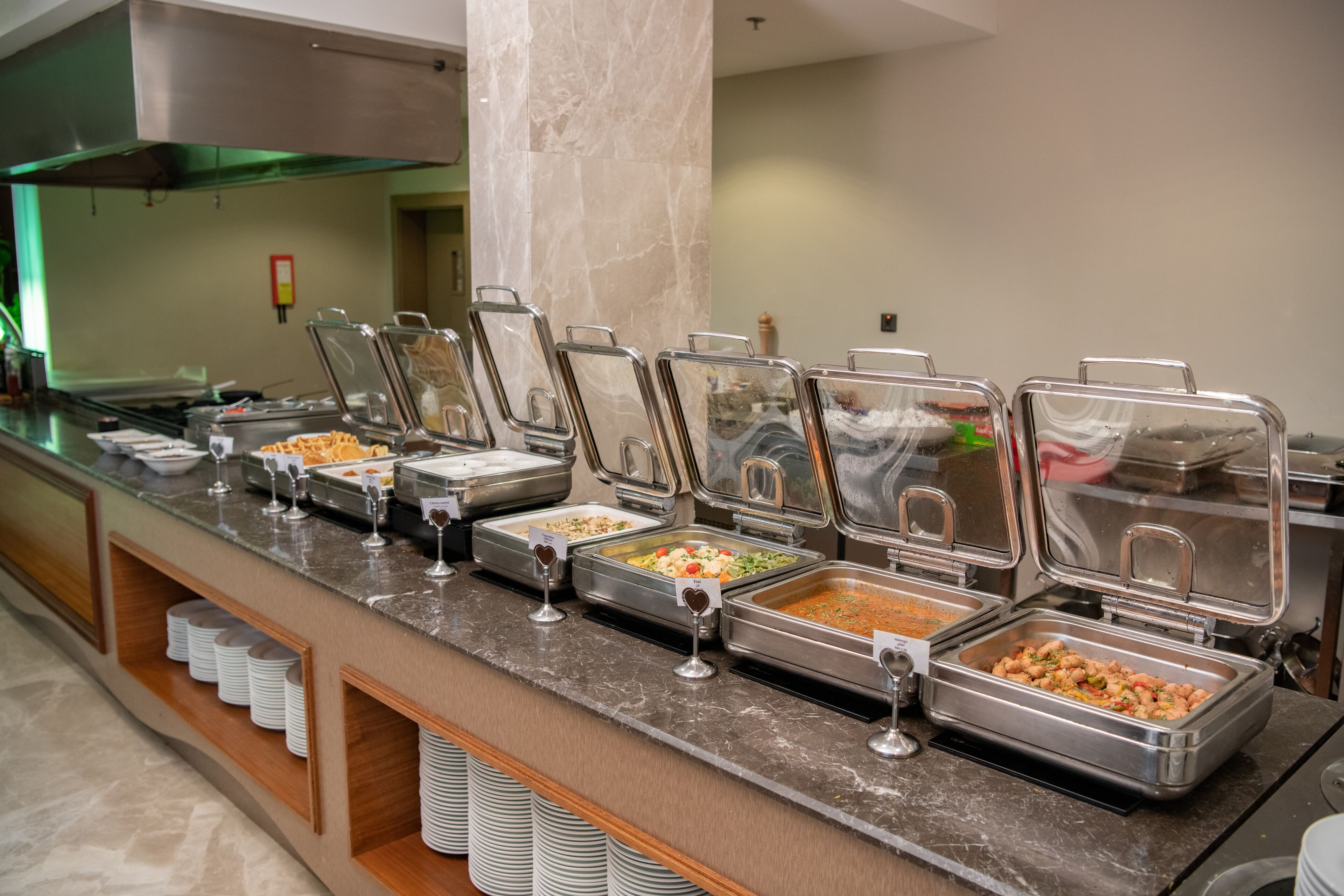 A buffet with various food options in metal containers, a marble countertop, and stacked white plates.