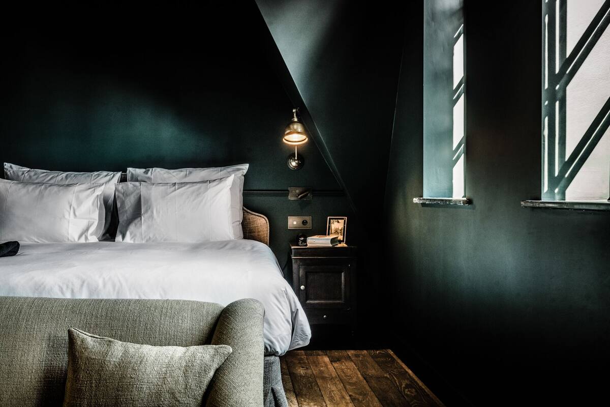 A bedroom with a bed, pillows, a nightstand, a wall-mounted lamp, and a window.