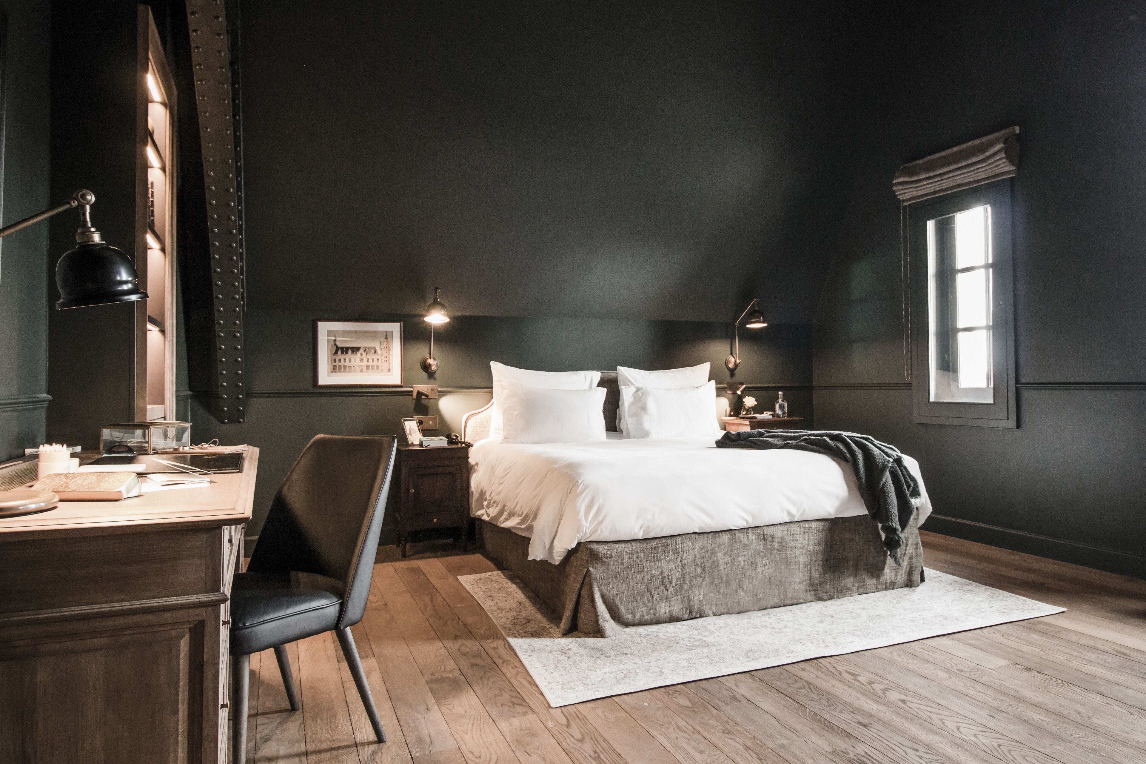 A bedroom with a large bed, a desk, and a chair. There is a window with curtains and a lamp on the desk.