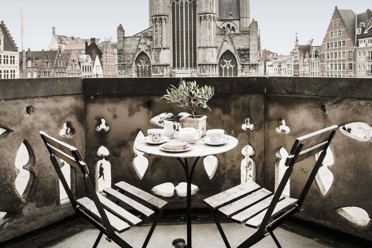 A table with chairs on a rooftop, overlooking a historic church with pointed towers.