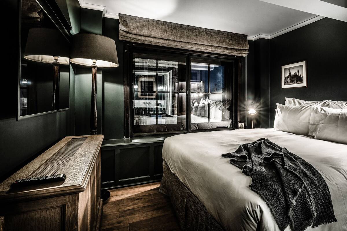 A modern hotel room with a large bed, a desk with a lamp, a window with a view of the city, and a framed picture on the wall.