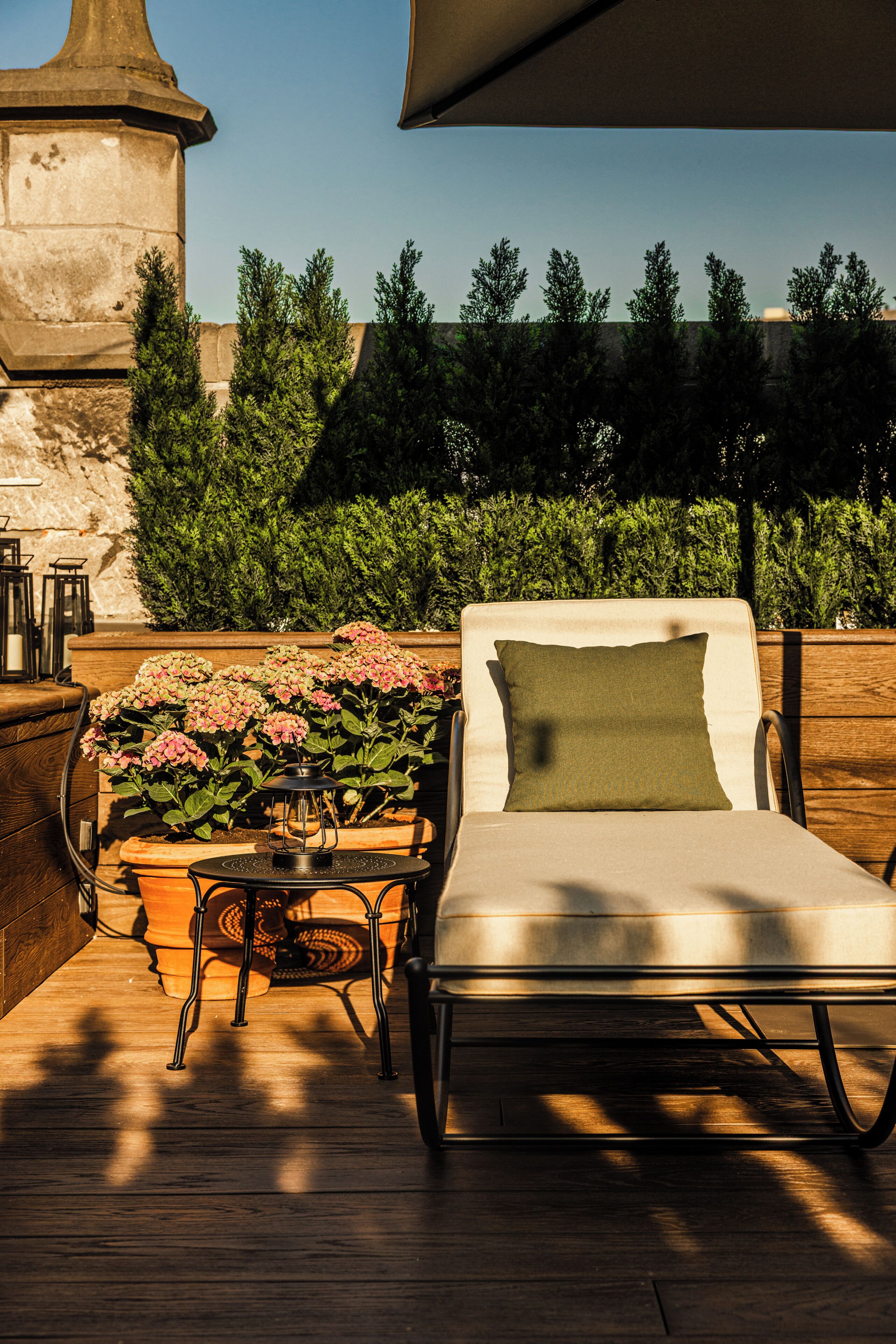A lounge chair with a cushion, a table with a lantern, and potted plants with pink flowers.