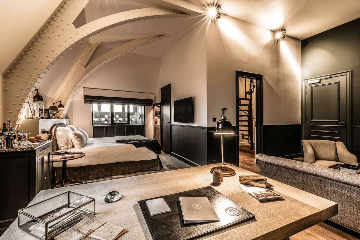A modern hotel room with a large bed, a desk with a lamp, a sofa, and a view of the city through the window.