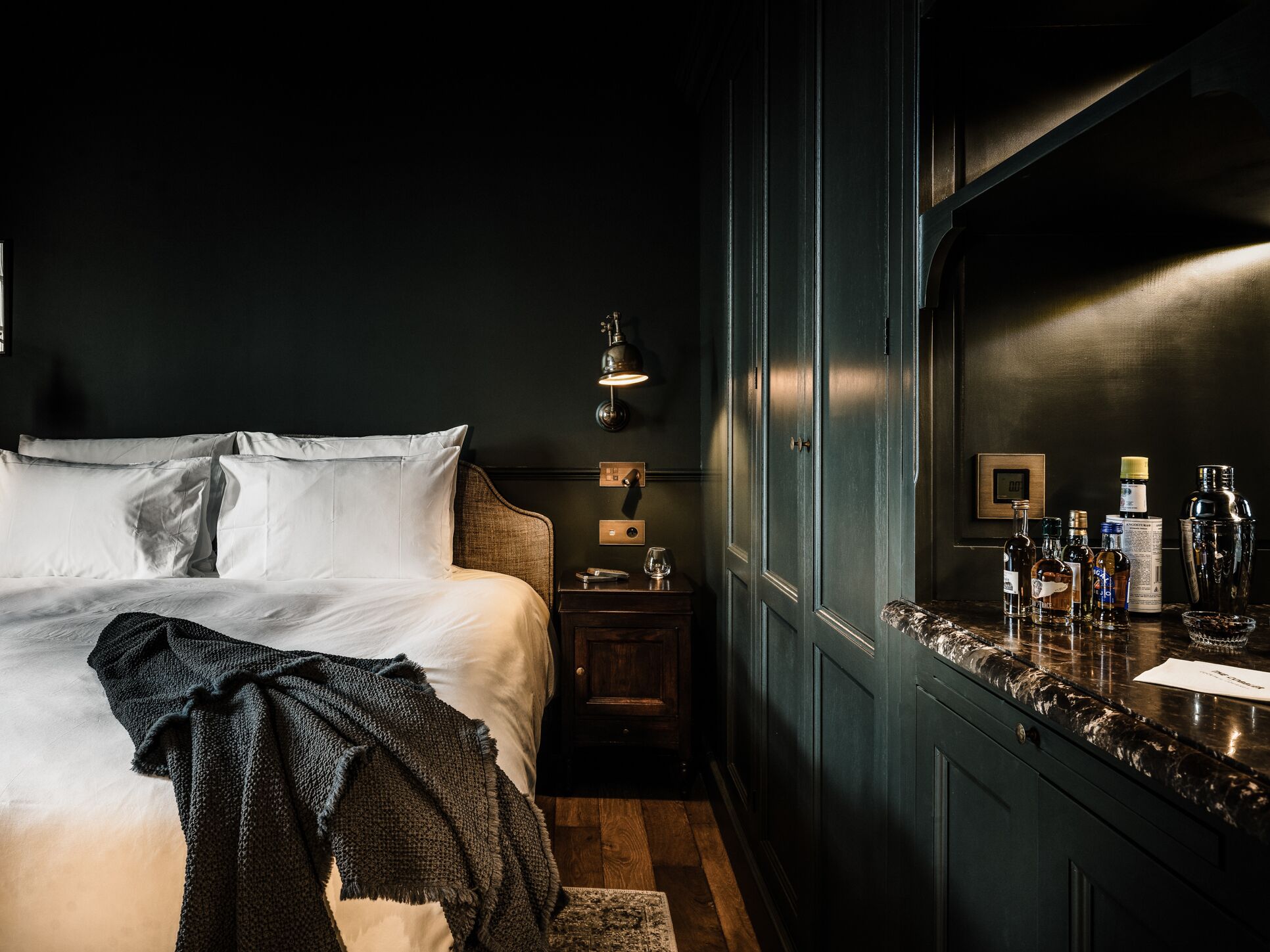 Ein schlafgemütliches Zimmer in dunklen Tönen mit einem großen Bett, Nachttischen und einem Spiegel mit verschiedenen Flaschen.