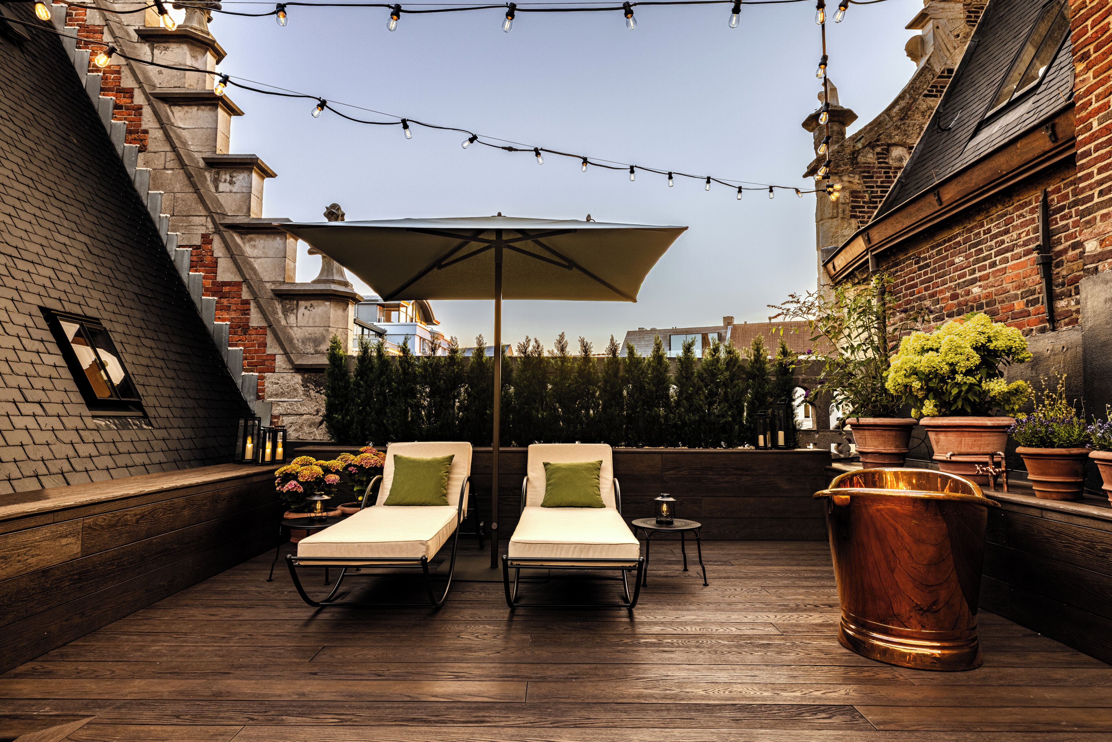 A rooftop patio with two lounge chairs, an umbrella, string lights, and potted plants.