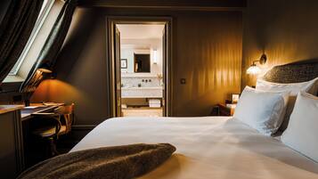 Ein Hotelzimmer mit einem großen Bett, einem Schreibtisch mit Stuhl, einem Fenster mit Vorhängen und einem durchgegangenen Türspalt sichtbaren Badezimmer.