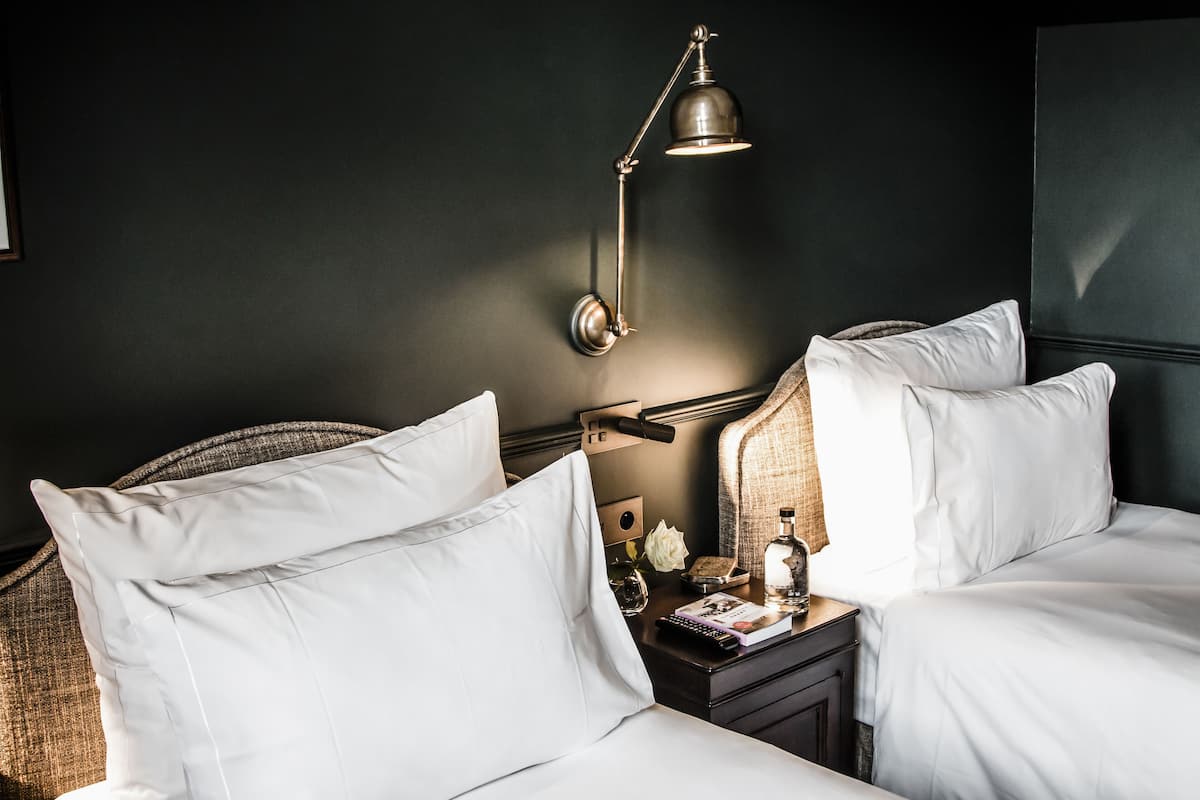 A hotel room with two beds, a bedside table with a lamp, a book, and a bottle.