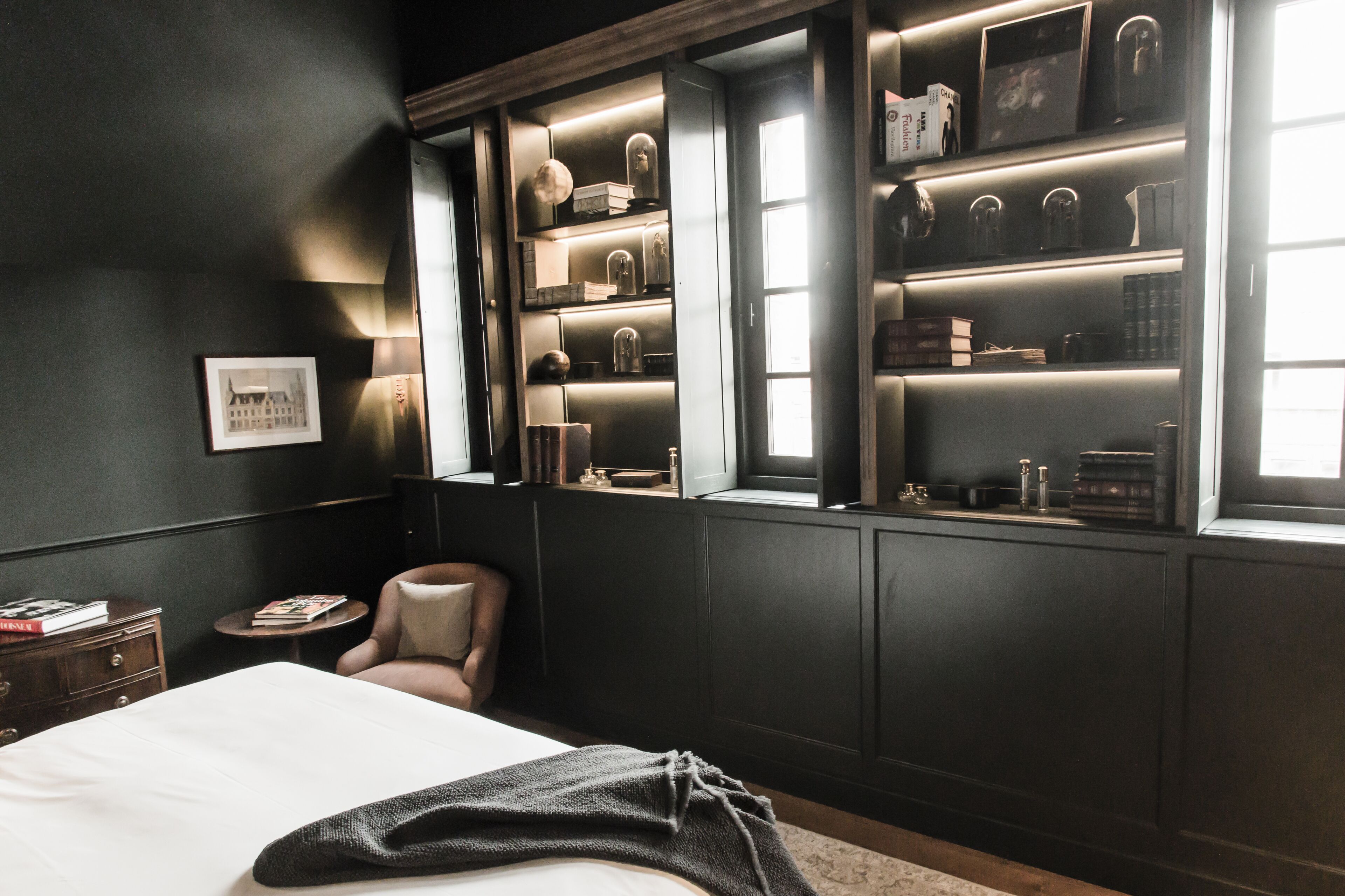 A bedroom with a large bed, a chair, a nightstand, and a built-in bookshelf with various items.