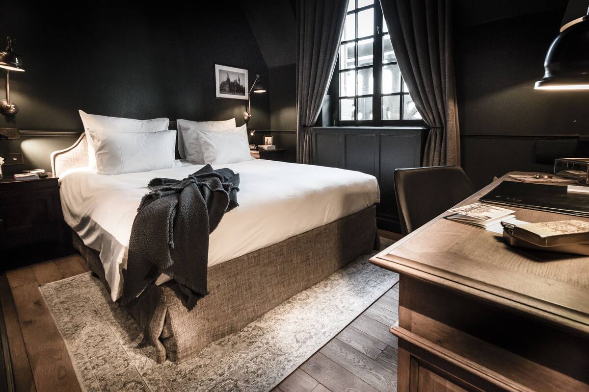 A bedroom with a large bed, a desk with a chair, a window with curtains, and a radiator.