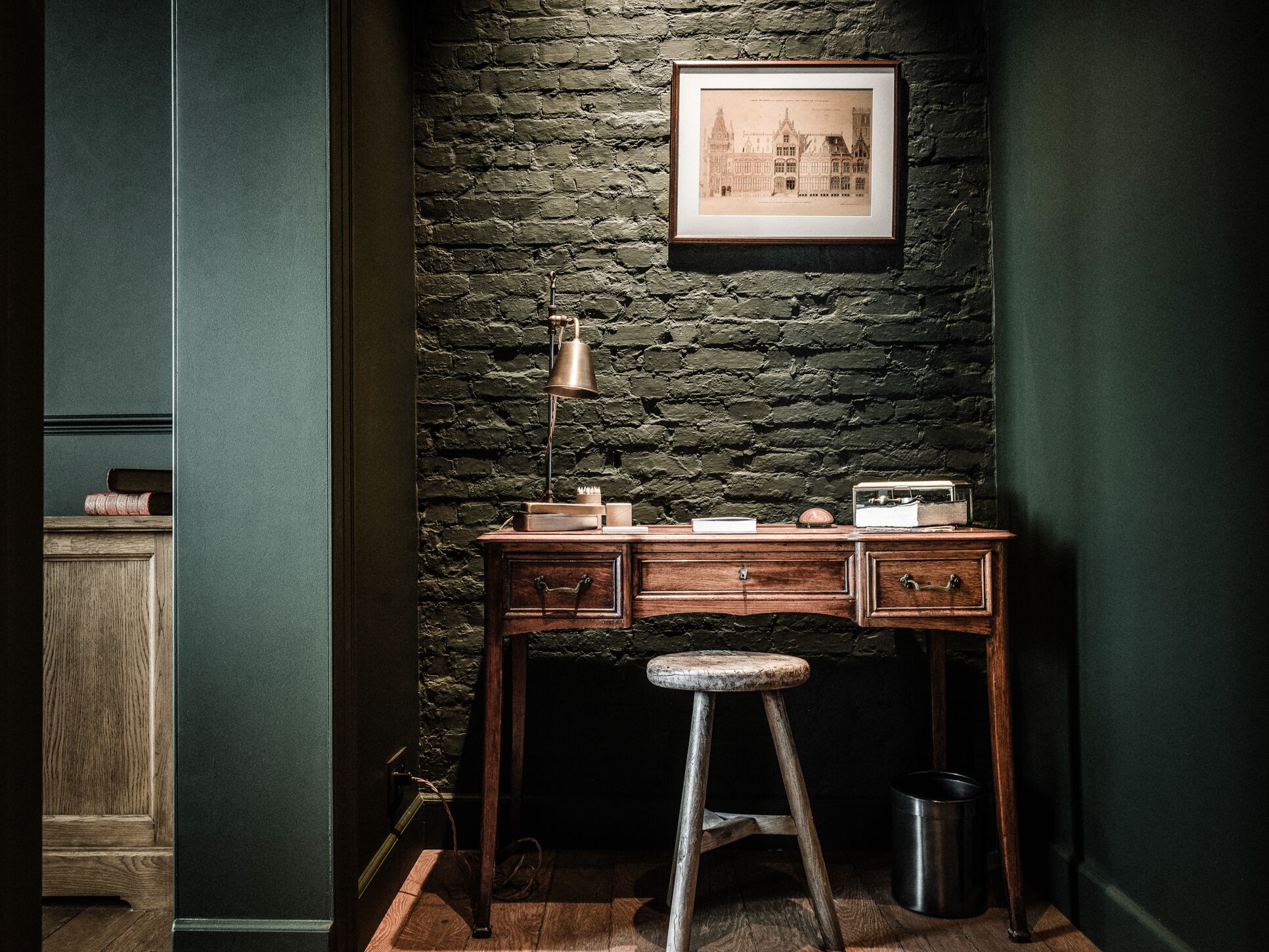 A room with a dark green wall, a wooden desk with drawers, a metal lamp, and a framed picture above the desk.