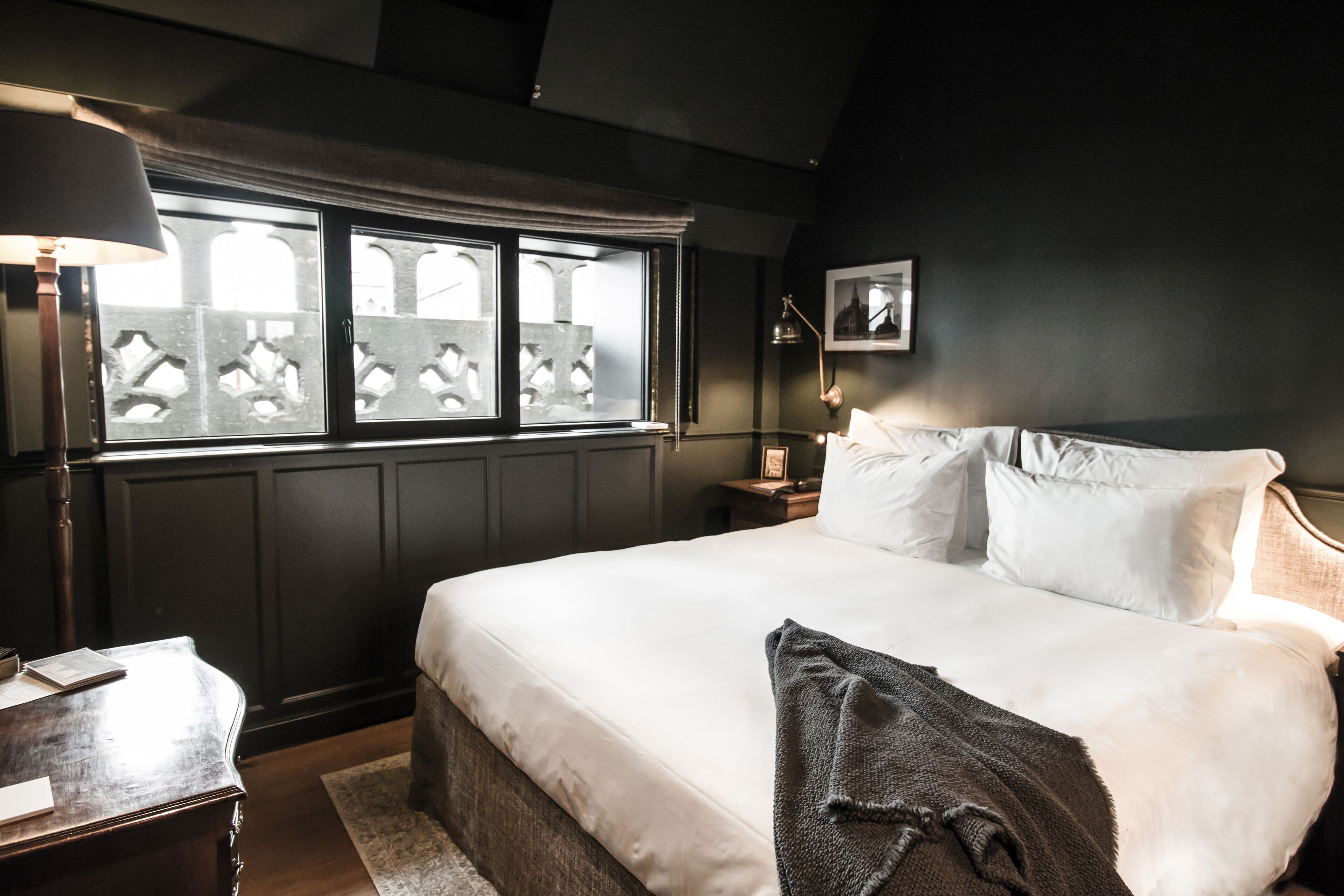 A bedroom with a large bed, a nightstand, a lamp, a window with a view, and a framed picture on the wall.
