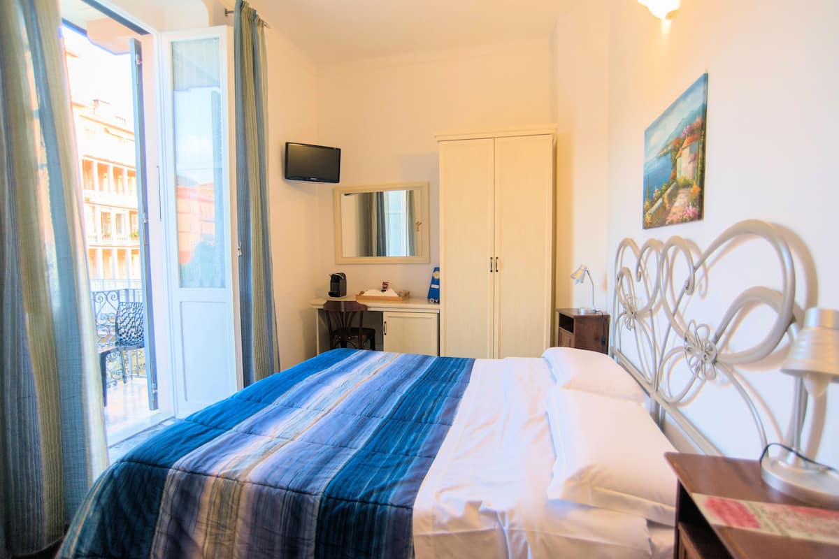 A hotel room with a bed, a desk with a chair, a wardrobe, a television, and a painting on the wall.