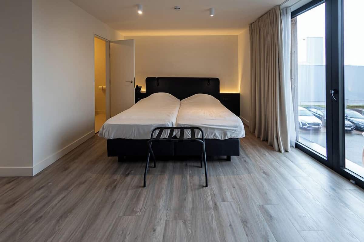 A modern hotel room with a large bed, a chair, and a view of parked cars through the window.