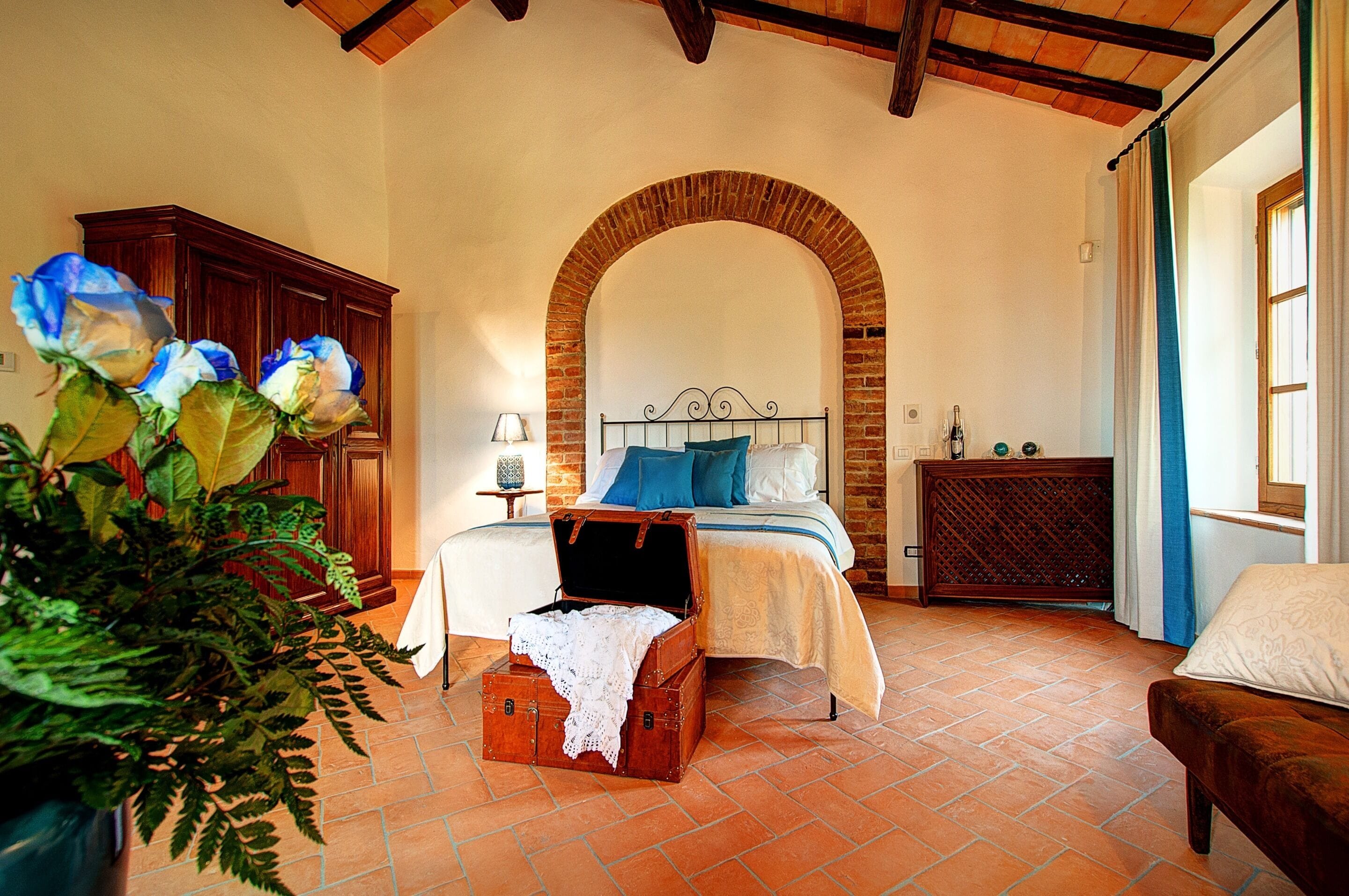 Un dormitorio con un arco de ladrillo, una cama con almohadas azules, un cofre de madera y una planta.