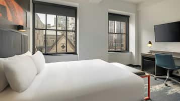 Ein Hotelzimmer mit einem großen Bett, einem Schreibtisch, einem Stuhl, einem Fernseher und zwei Fenstern mit Blick auf die Stadt.