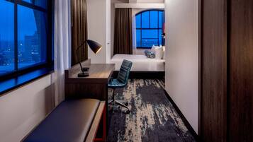 Ein Hotelzimmer mit einem Bett, einem Schreibtisch, einem Stuhl, einer Bank und einem großen Fenster mit Blick auf die Stadt.