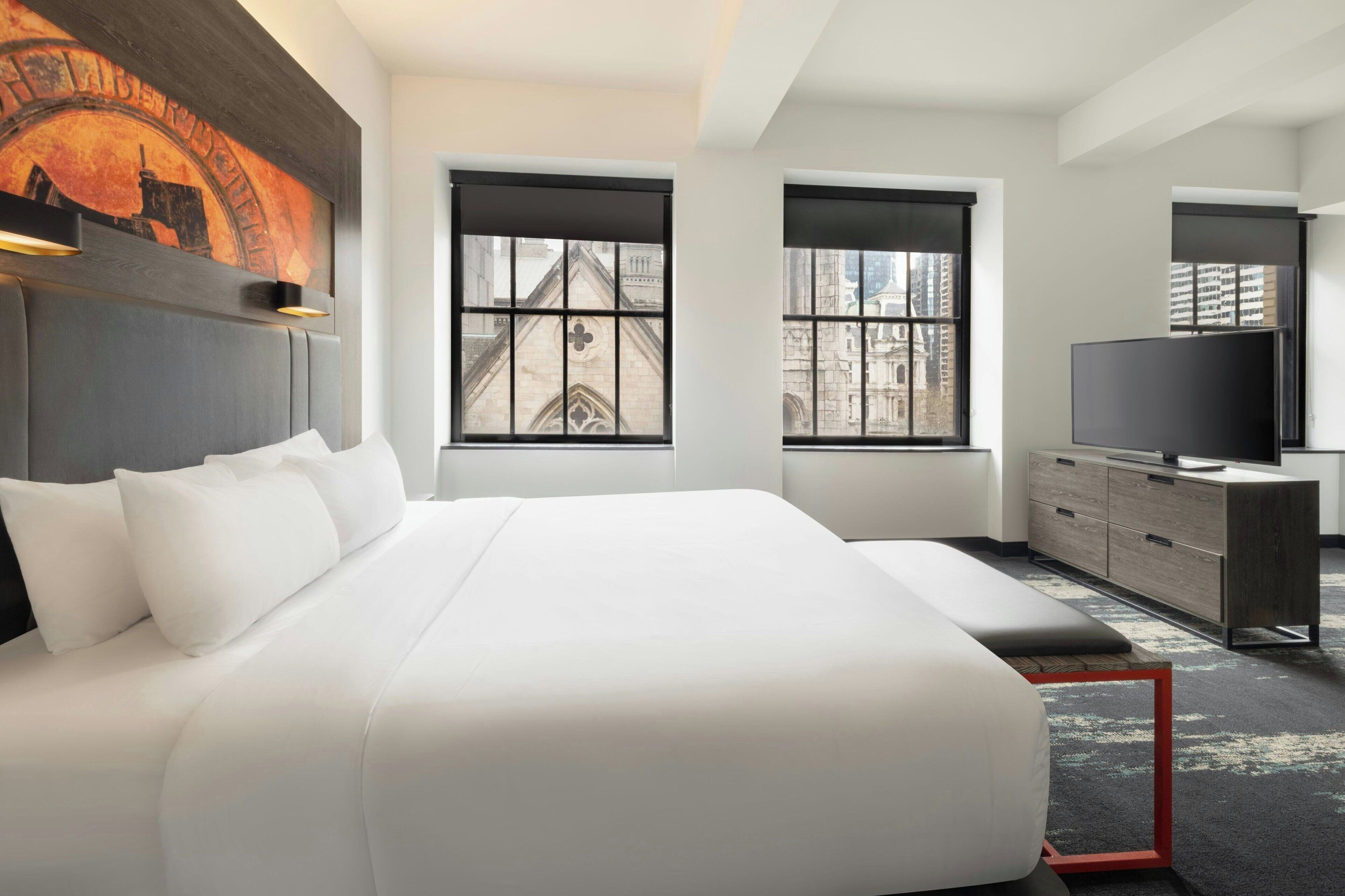 A modern hotel room with a large bed, two windows, a television, and a desk.