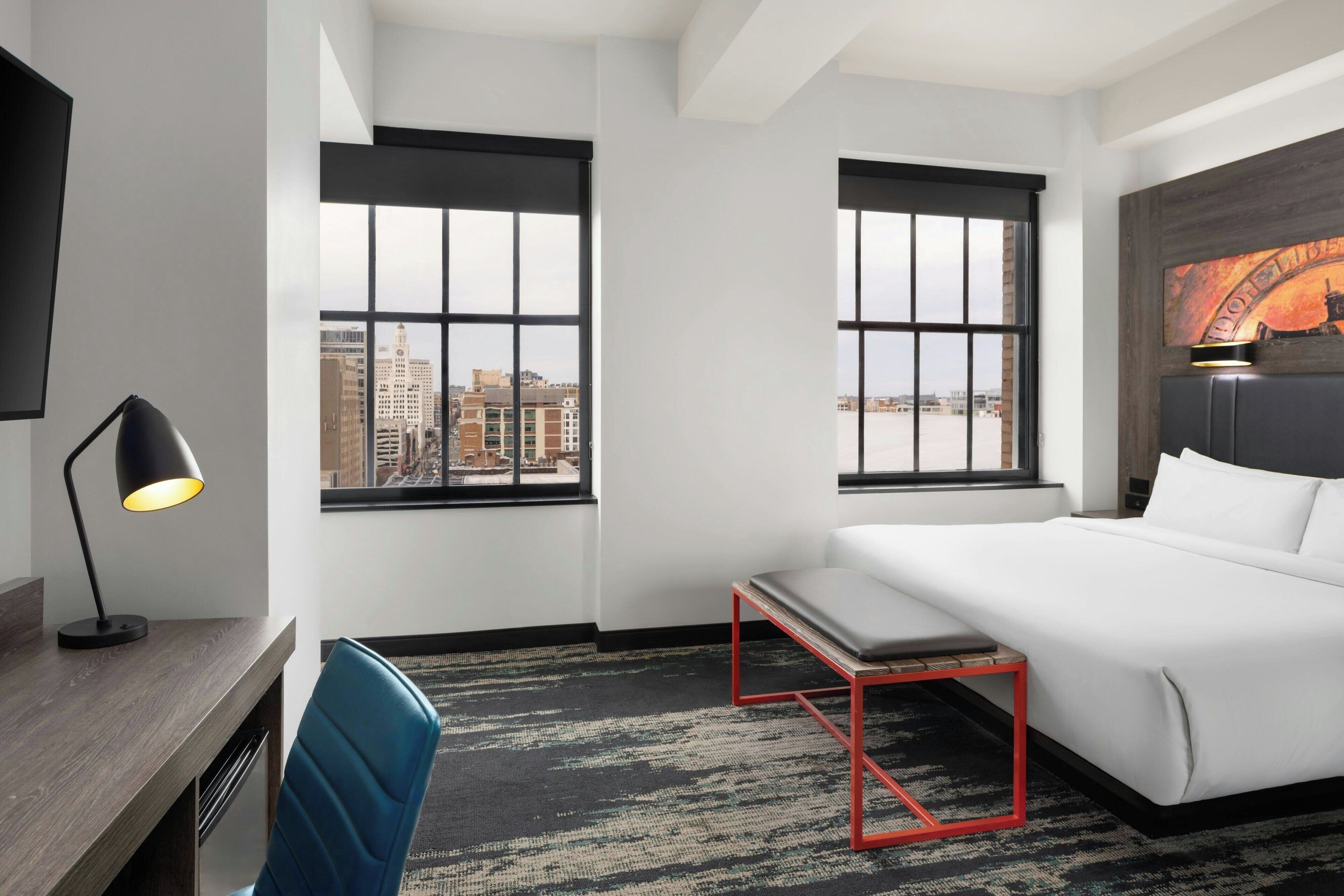 Habitación de hotel con cama, escritorio, silla, lámpara de escritorio y dos ventanas con vistas a la ciudad.