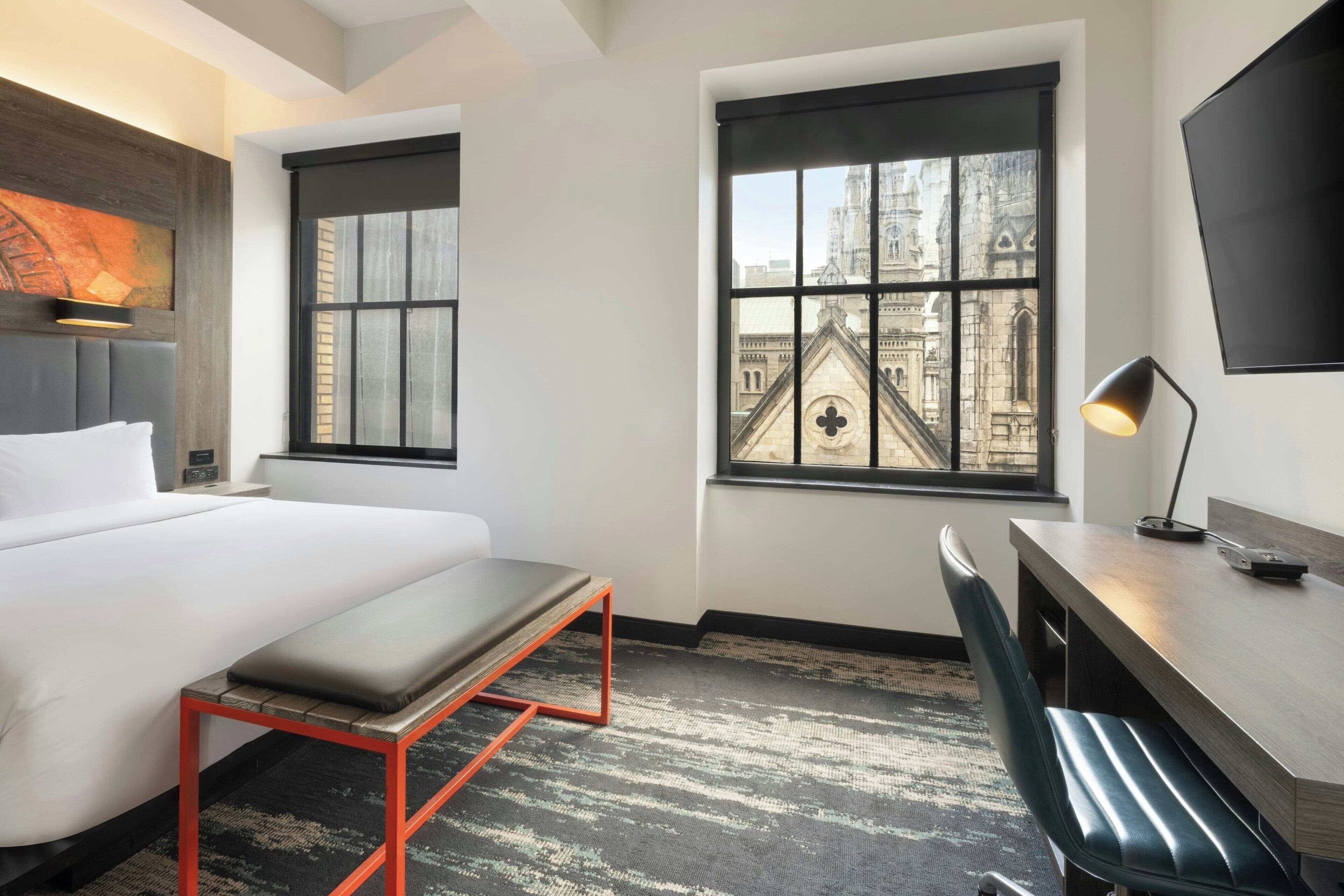 A hotel room with a bed, a desk, a bench, and two windows offering views of a historic building.
