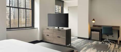 Une chambre d’hôtel avec un lit, un téléviseur posé sur une commode en bois, un bureau avec une chaise et une grande fenêtre donnant sur la ville.