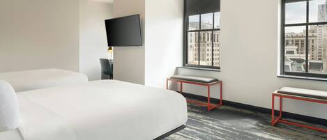 A modern hotel room with a large bed, a desk, a chair, a TV, and a window with a city view.
