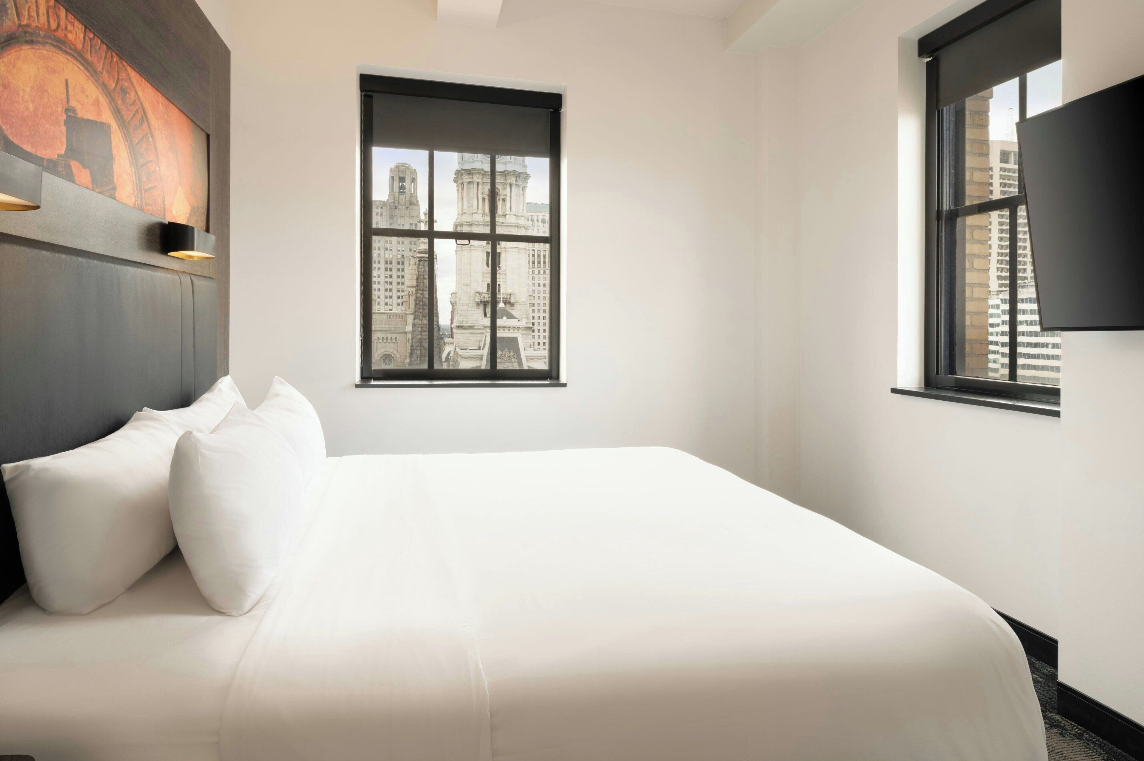 A hotel room with a large bed, a window with a cityscape view, and a mounted TV on the wall.
