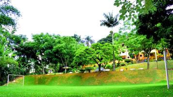 Um parque verdejante com um gol de futebol, árvores e um edifício com fachada amarela.