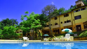 Una piscina con sillas reclinables blancas y sombrillas, rodeada de árboles y un edificio con balcones.